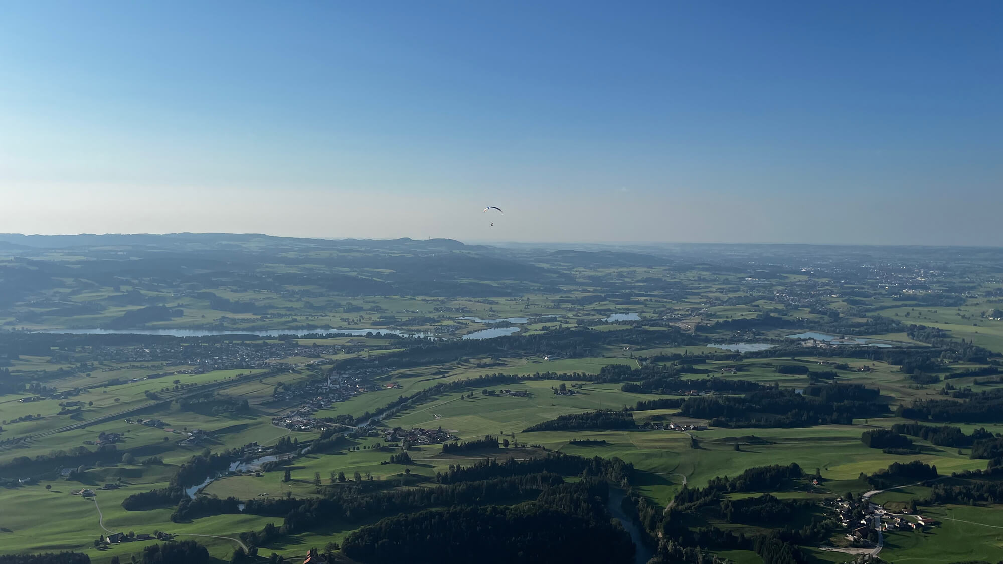 Luftbild über eine weite hügelige grüne Landschaft mit Wäldern und Flüssen, darüber blauer Himmel