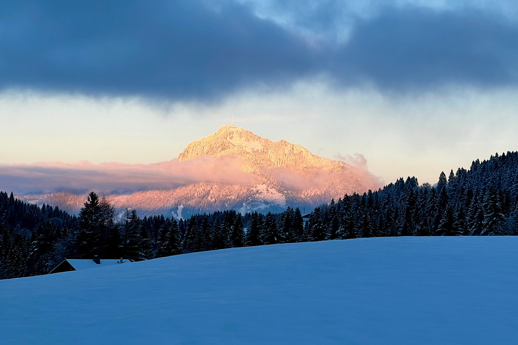 Wolkenverhangene dunkle Schneelandschaft, in der Ferne leuchtet ein schneebedeckter Berg von der Sonne beschienen in Orange- und Rosatönen