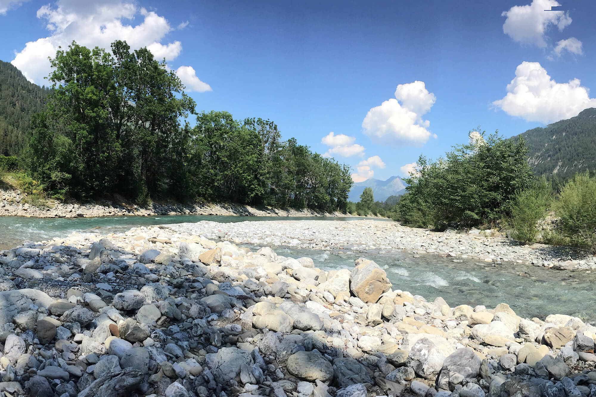 Ein Gebirgsbach schläbgelt sich über große weiße Steine, links und rechts dunkelgrüne Büsche, blauer Himmel mit Schäfchenwolken